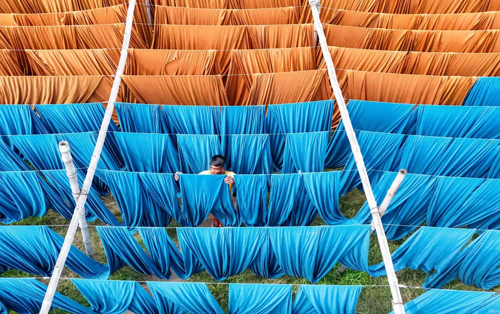 Man hangs up blue cloth on a line to dry. He is surrounded by similar blue and orange cloths. 