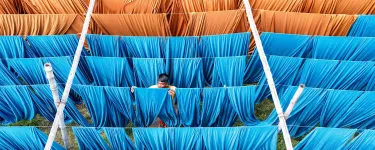 Man hangs up blue cloth on a line to dry. He is surrounded by similar blue and orange cloths. 